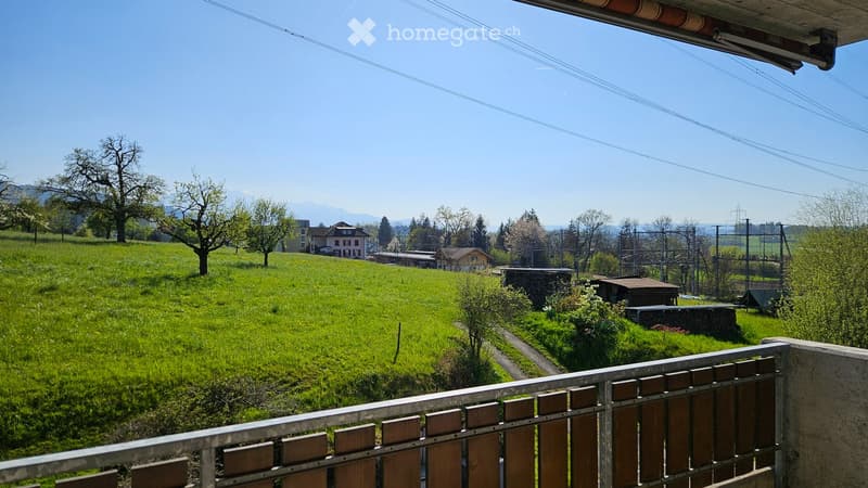 View from a balcony or terrace overlooking a green, hilly landscape with scattered trees, houses, and distant mountains under a clear blue sky.
