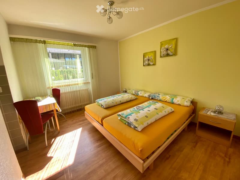 Bright bedroom with two single beds, a small table with a chair, and a nightstand with a lamp. Yellow walls, wooden flooring, and large window with curtains.