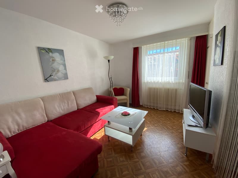 Living room with a red sectional sofa, a glass coffee table, a single armchair, a TV on a stand, a floor lamp, and artwork on the walls.