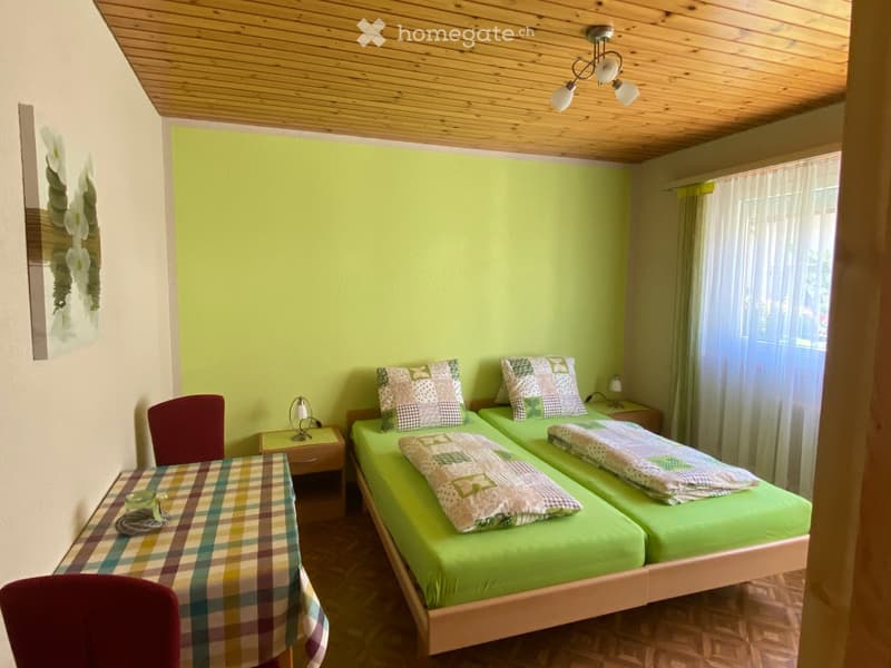 Cozy bedroom with two single beds, green bedding, nightstands, a small table with chairs, and a decorative wall piece.