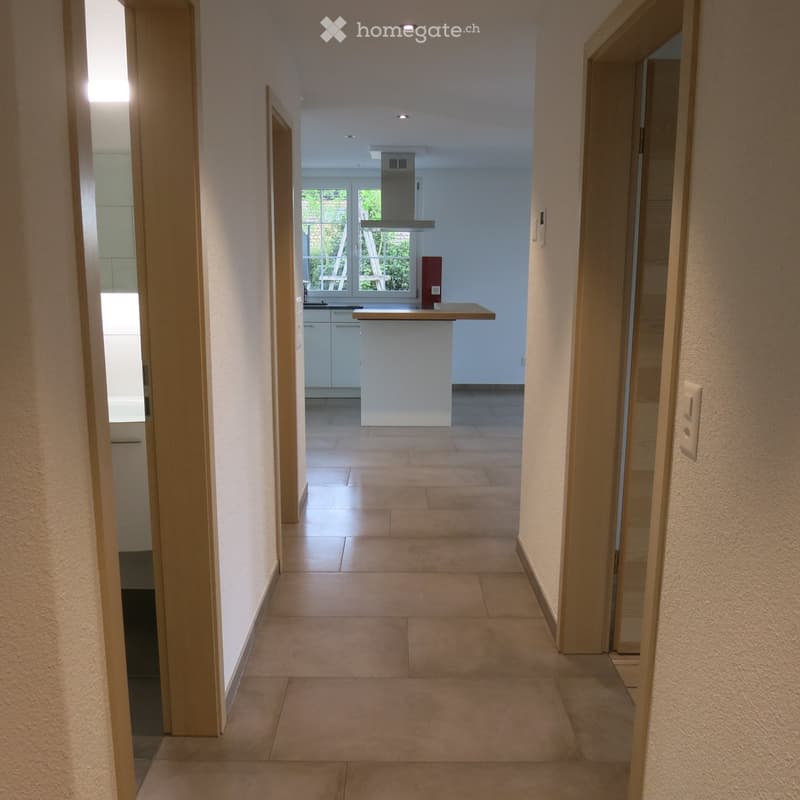 Empty hallway with tiled floor, light-colored walls, and a view into a modern kitchen area with white cabinets, a central island, and large windows.