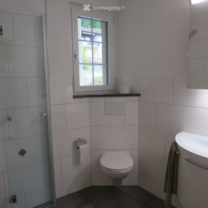 Modern bathroom with white tiles, a toilet, a sink, a shower, and a towel hanging on a rack. A window provides natural light.