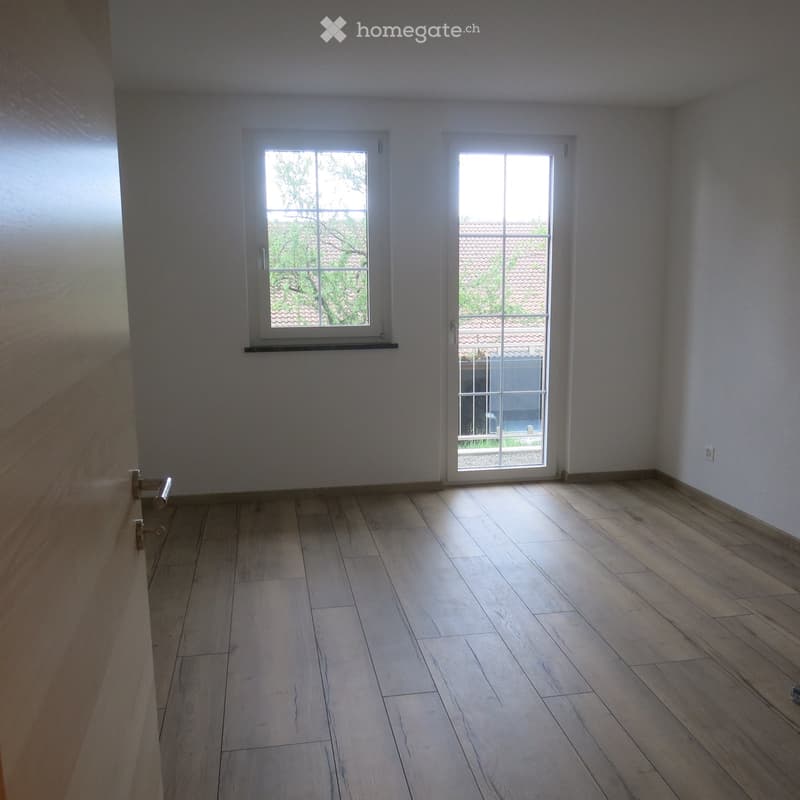 Empty room with light wooden flooring, white walls, and two windows, one of which is a sliding glass door leading outside.
