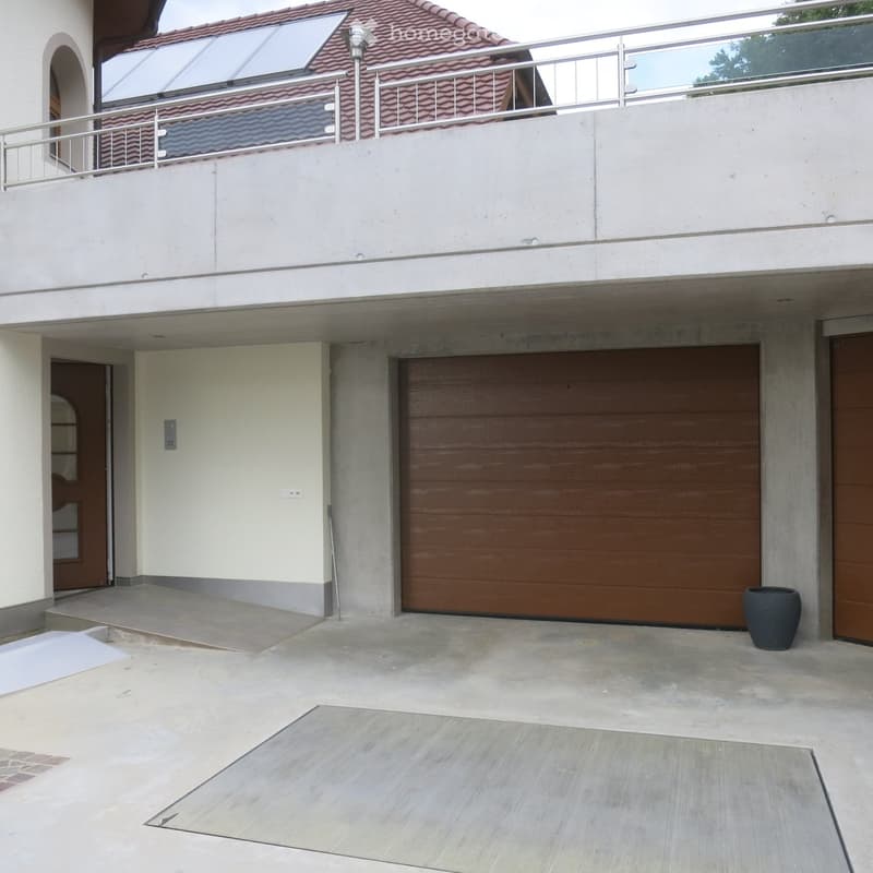 Modern two-story house with a garage, raised ground floor, and balcony with metal railing.
