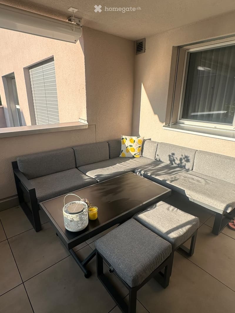 Balcony with grey sectional sofa, coffee table, two stools, decorative pillow, candle holder, and a cup.