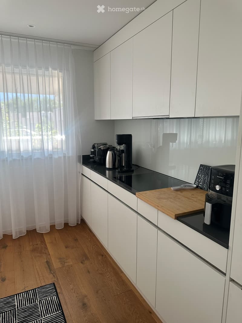 Modern kitchen with white cabinets, black countertops, wooden cutting board, coffee maker, oven, and a small rug on the floor.
