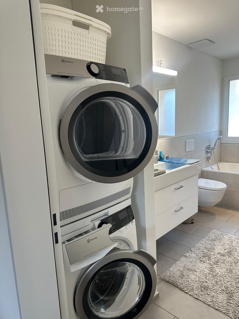 Modern bathroom with a stacked washer and dryer unit, a sink with a mirror, a toilet, a bathtub, and a shower area. The space features light-colored tiles and a small white rug on the floor.