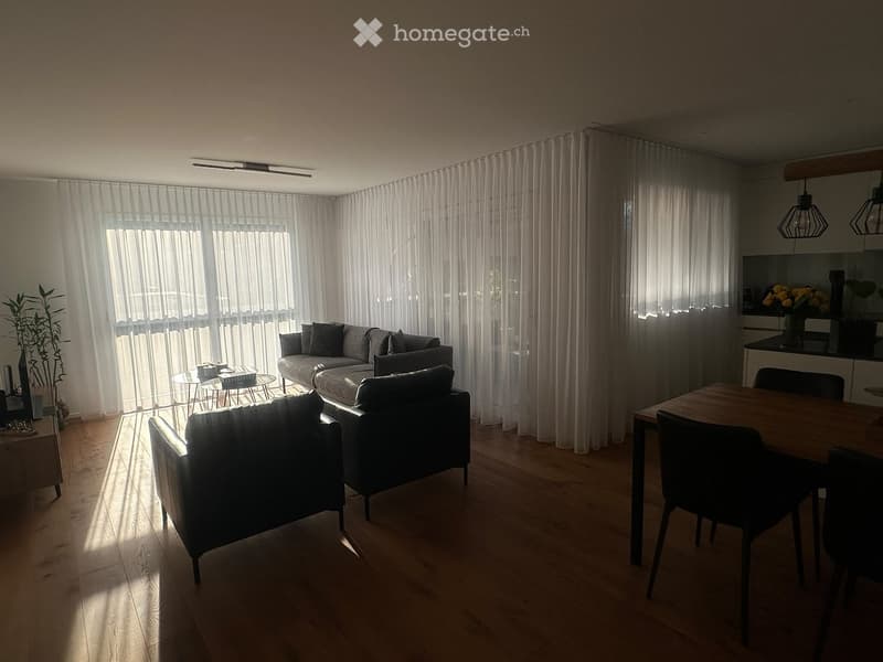 Bright living room with large windows covered by sheer curtains, a grey sofa, a coffee table, two armchairs, a dining table with chairs, and pendant lights.