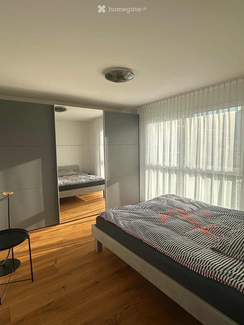 Modern bedroom with wooden flooring, large bed with striped duvet, sleek wardrobe with mirrored doors, and a small round table with a lamp.