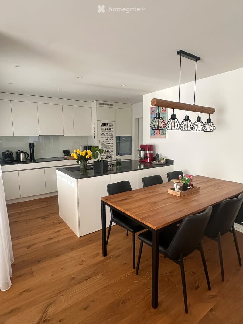 Modern kitchen with white cabinets, wooden dining table, black chairs, and various appliances including a coffee maker and blender. The space features a wooden floor and pendant lighting.