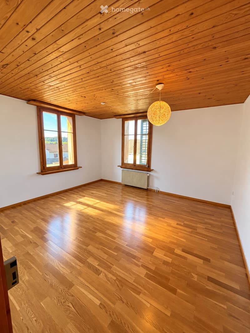 Empty room with wooden flooring, white walls, wooden ceiling, and large windows letting in natural light. A radiator is visible under one window, and a hanging light fixture is present.