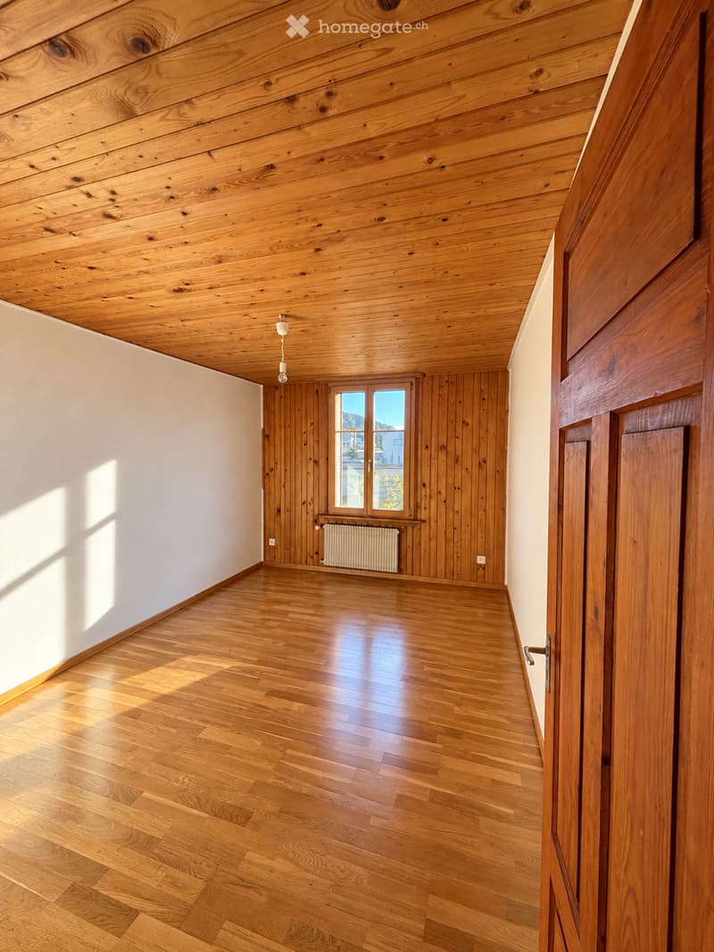 Empty room with wooden floors, wooden walls, and a large window with a view. The room features a simple, modern design with natural wood finishes and a single light fixture hanging from the ceiling.