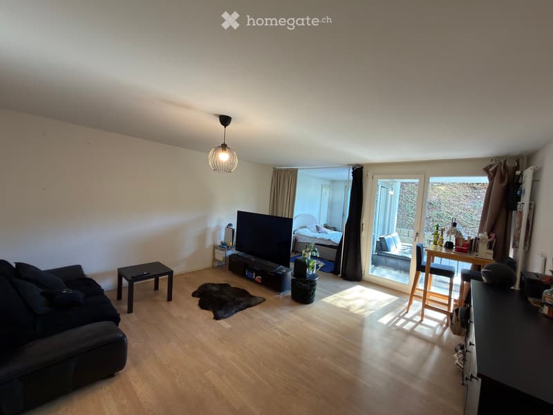 A modern living room with a dark blue sofa, coffee table, television, and a small table with chairs. The room features wooden flooring, a hanging lamp, and a view of a bedroom through an open doorway.