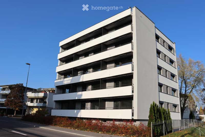 Modern multi-story residential building with white facade, multiple balconies, and surrounding greenery.