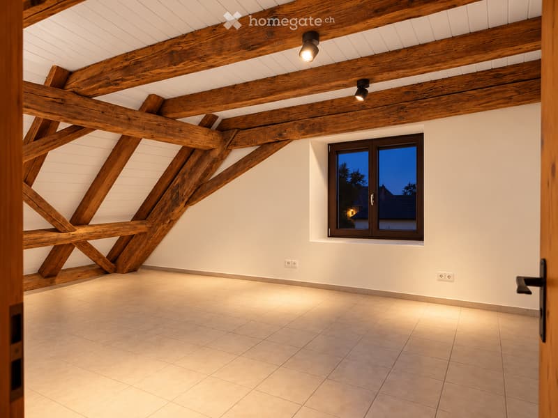 Empty attic space with exposed wooden beams, tiled floor, and a single window showing a view of the evening sky.
