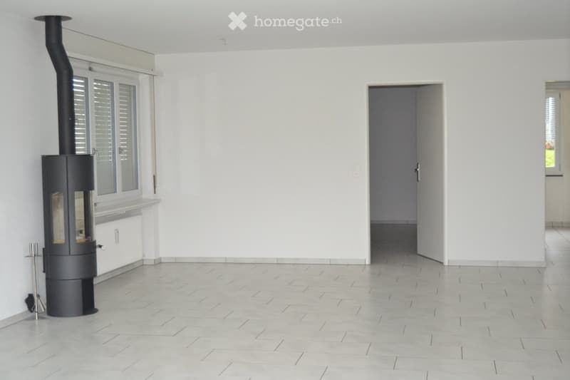 Empty room with light-colored tiled flooring, white walls, and a modern black wood-burning stove with a chimney. There is a doorway leading to another room.