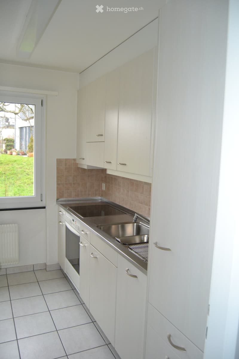 Modern kitchen with white cabinets, stainless steel sink, and tiled floor. Features a window with a view of a garden.