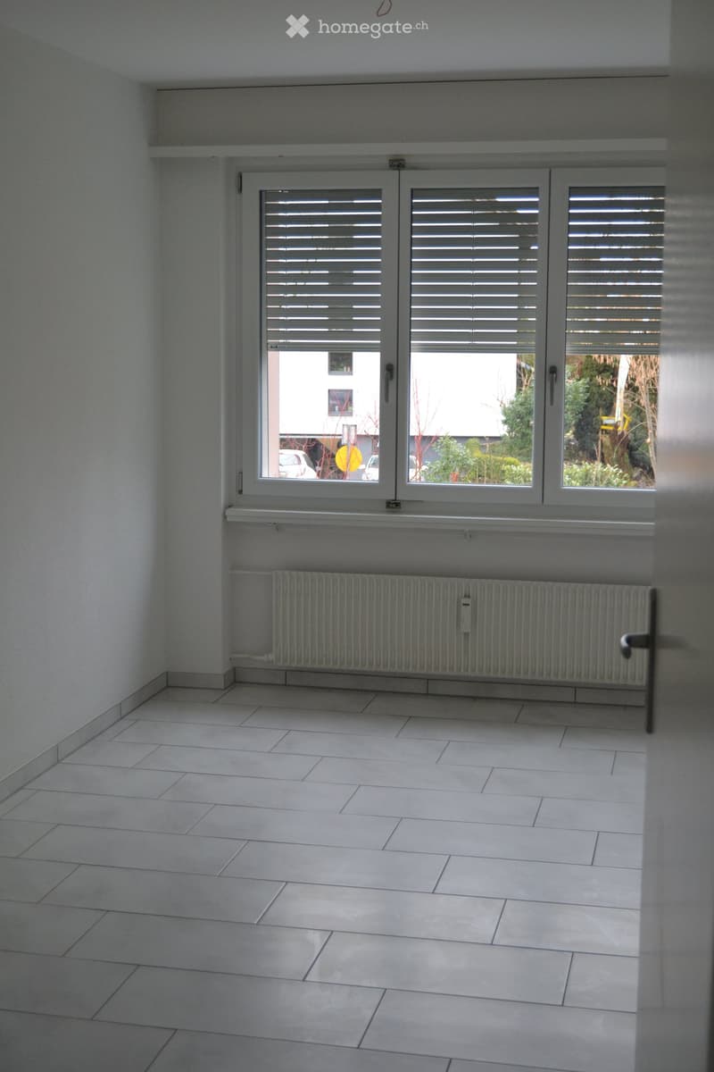 Empty room with white tiled flooring, white walls, large window with blinds, radiator, and a door handle visible.