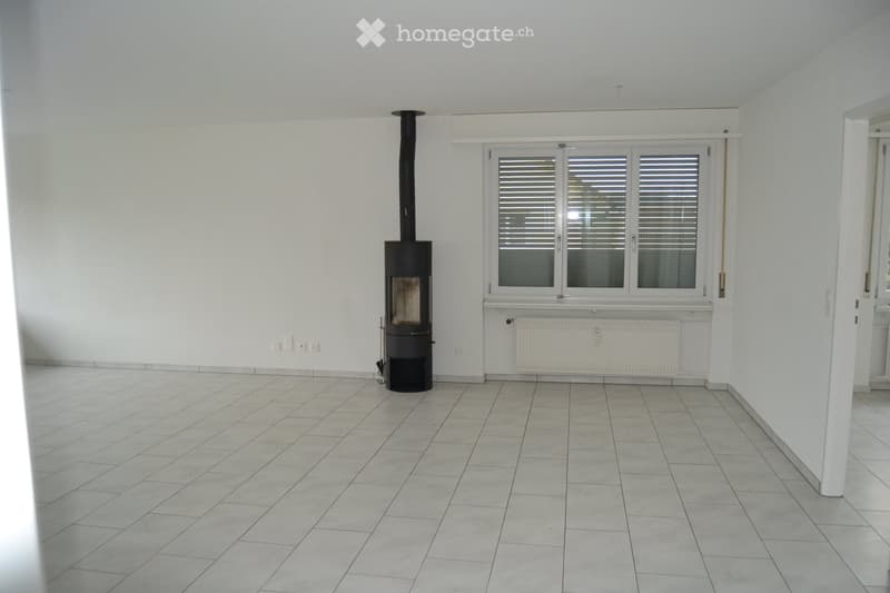 Empty room with tiled floor, white walls, large window with blinds, and a black wood-burning stove with chimney.