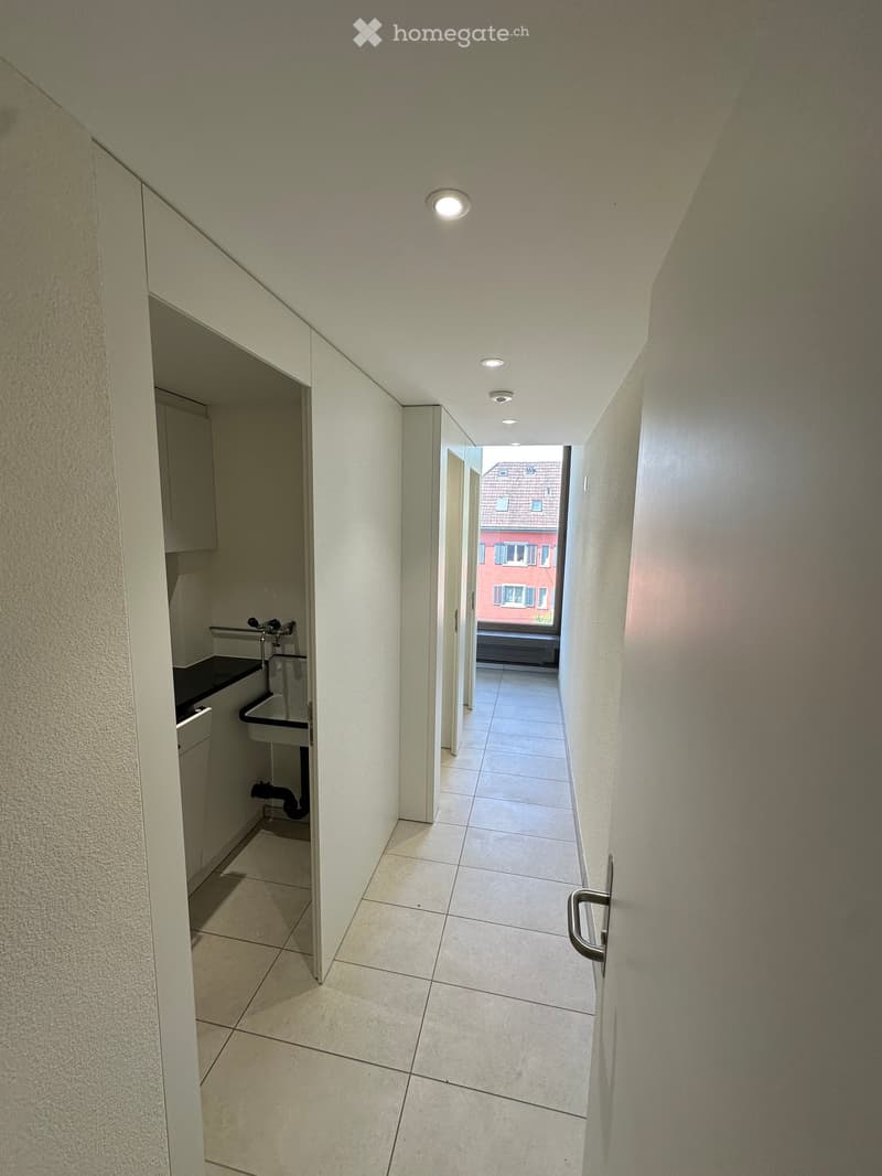 A narrow hallway with tiled flooring, white walls, and recessed ceiling lights. A small sink area is visible on the left, and a door leads to an outdoor area with a view of a red building.