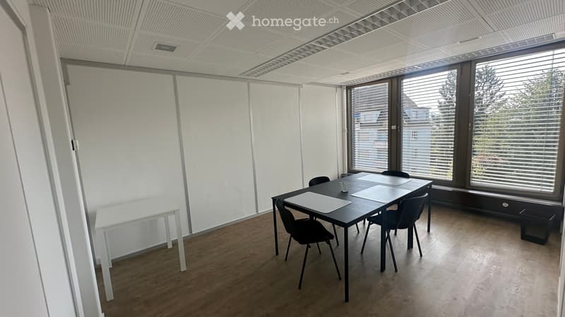 Bright, modern room with large windows, white walls, and a wooden floor. Contains a black table with chairs, a white side table, and a cup on the table.