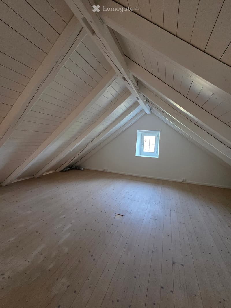 An attic space with wooden plank flooring and exposed wooden beams on the ceiling. A small window is positioned near the peak of the roof, allowing natural light to enter. The room is completely empty, with no furniture or decorations.