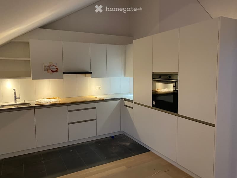 Modern kitchen with white cabinets, stainless steel sink, and built-in oven. The space features a mix of dark tiled and wooden flooring.