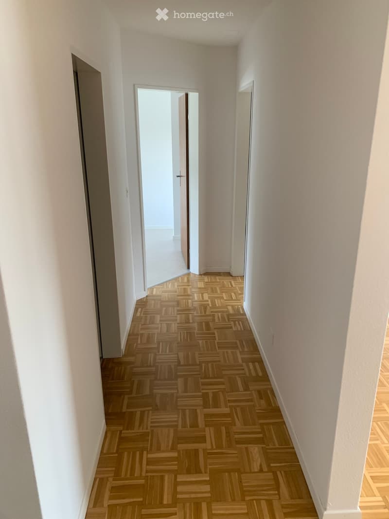Empty hallway with wooden herringbone flooring and white walls, featuring two open doors at the end.