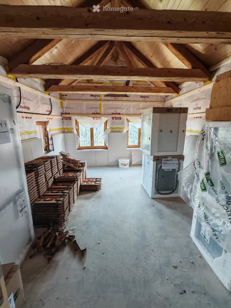 Empty attic space with exposed wooden beams, insulation, and construction materials. Windows at the far end, stacks of wooden planks, and a bucket on the floor.