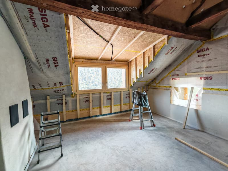 An attic space under construction with exposed wooden beams, insulation, and unfinished walls. There are two ladders, a small window, and construction materials visible.