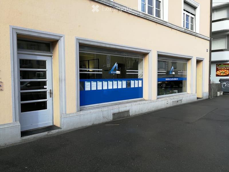 Two-story building with a light yellow facade, featuring a garage door on the left and a storefront with blue signage on the right.