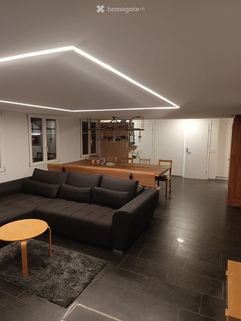 Modern living and dining area with dark tiled flooring, a sectional sofa, a small round wooden stool, a dining table with chairs, and a hanging light fixture.