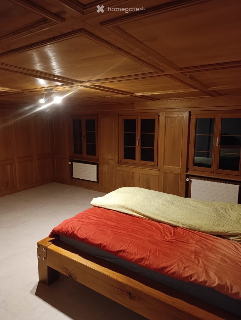 Cozy wooden bedroom with a single bed, red and yellow blankets, wooden walls, and built-in wooden cabinets with glass doors.