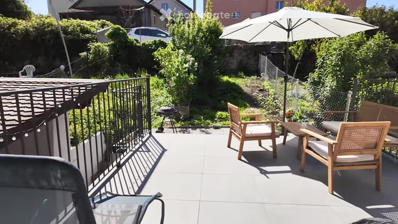 Balcony with wooden chairs, a table, and an umbrella, surrounded by greenery and a fence.