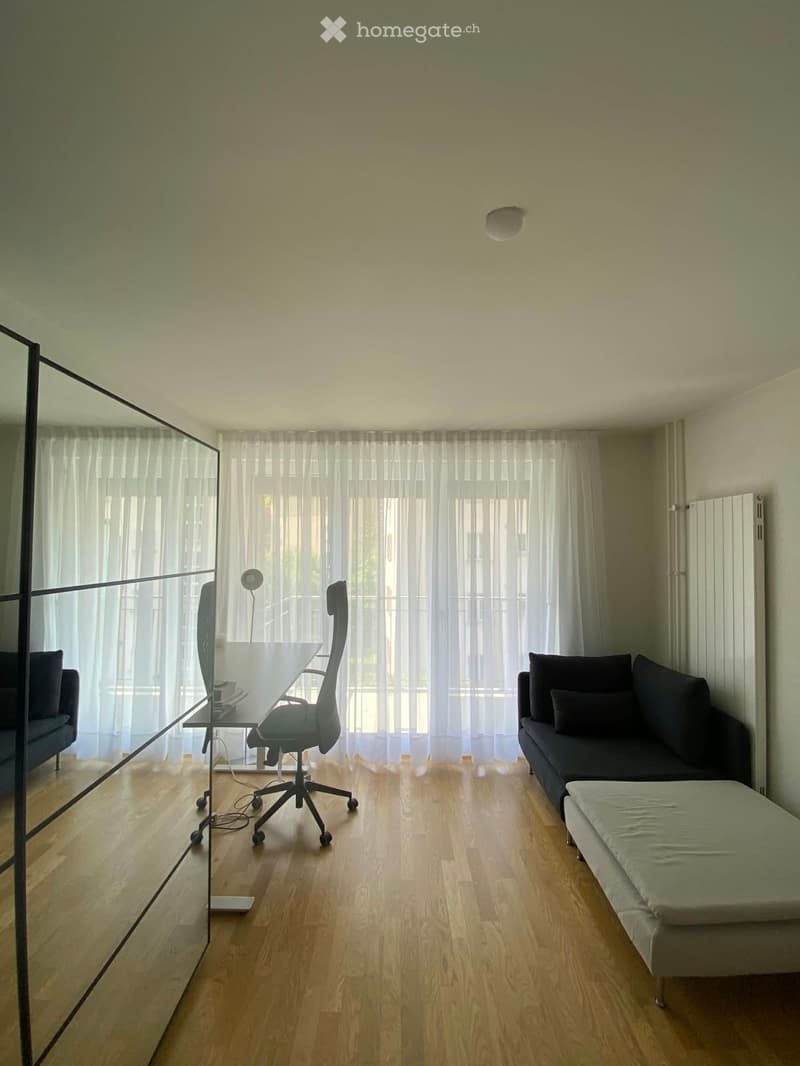 A modern, open-concept living and working space with a sofa, ottoman, desk, office chair, and a large mirror. The room features light wooden flooring and large windows with sheer curtains.