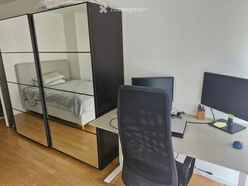 A modern, well-lit room with a desk, chair, laptop, and large mirror reflecting a bed with pillows and a blanket. The room features wooden flooring and a sleek, contemporary design.