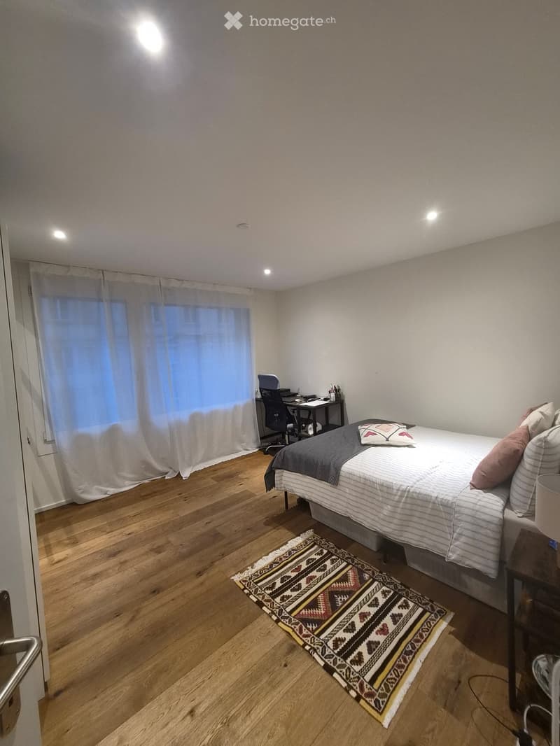 A small, well-lit bedroom with a wooden floor, a bed with striped bedding, a patterned rug, a desk with a chair, and a window with sheer curtains.