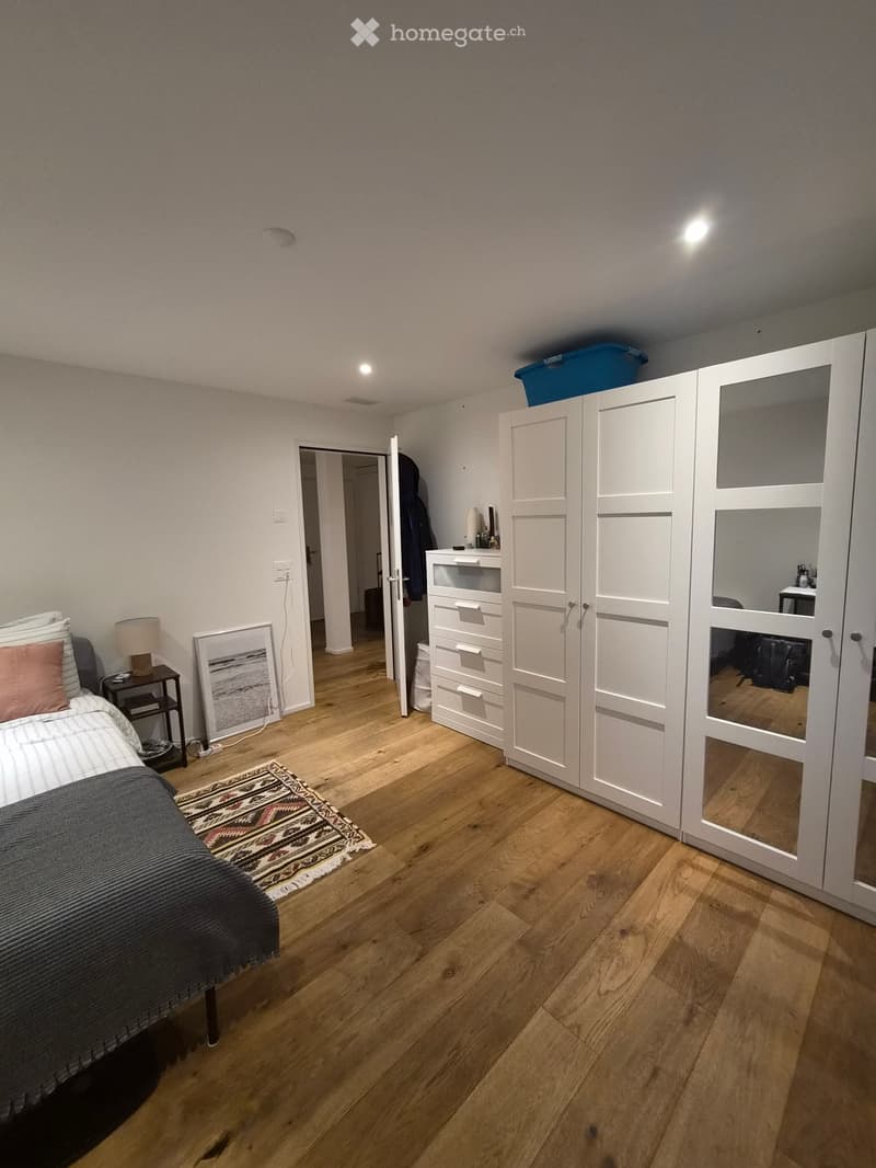 Cozy bedroom with wooden flooring, a neatly made bed with pillows and a blanket, a nightstand with a lamp, a picture frame on the floor, a patterned rug, and a large white wardrobe with mirrored doors.
