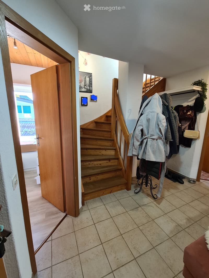 Hallway with tiled floor, wooden staircase, coat rack with jackets and bags, and an open door leading to another room.