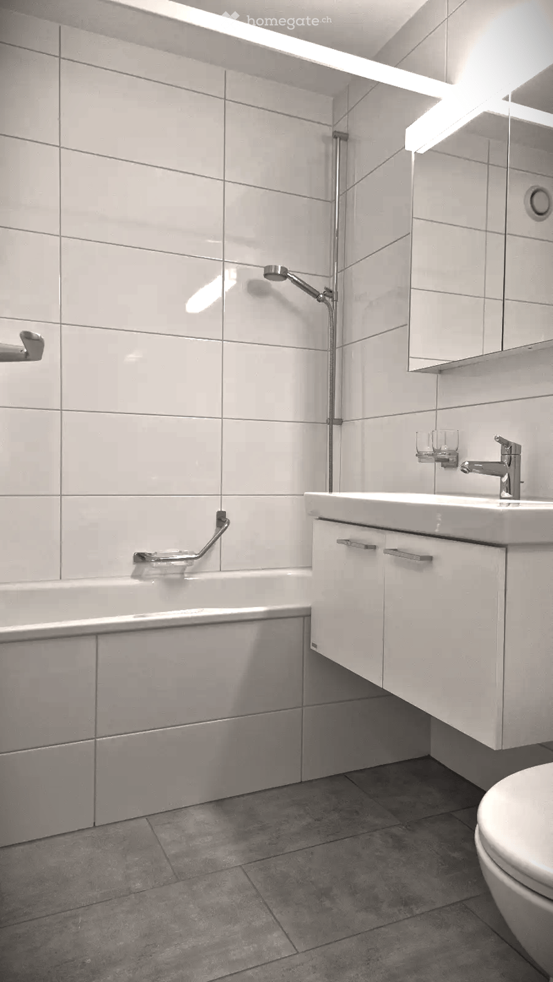 Modern bathroom with a bathtub, sink, mirror, and toilet. Features grey tiles on walls and floor, sleek cabinetry, and a modern faucet.
