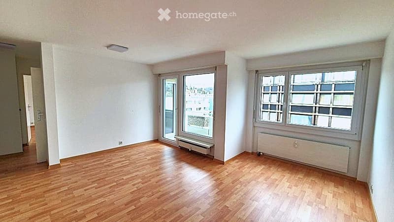 Empty room with wooden flooring, white walls, large windows, and a sliding glass door leading to a balcony.
