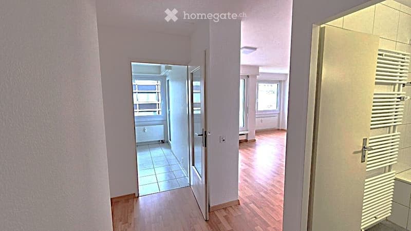 A clean, well-lit hallway with wooden flooring, white walls, and several doors leading to different rooms. The hallway features a glass door on one side and a radiator on the other.