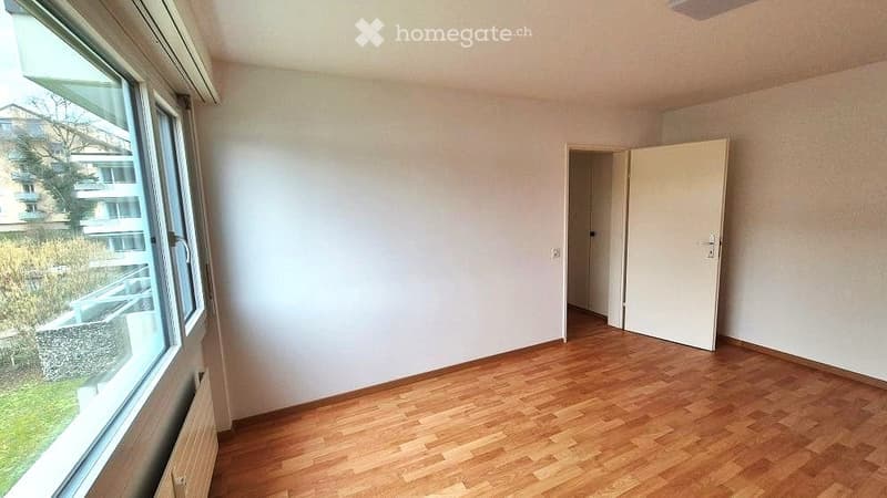 Empty room with wooden flooring, white walls, large window with view, and an open doorway leading to another space.