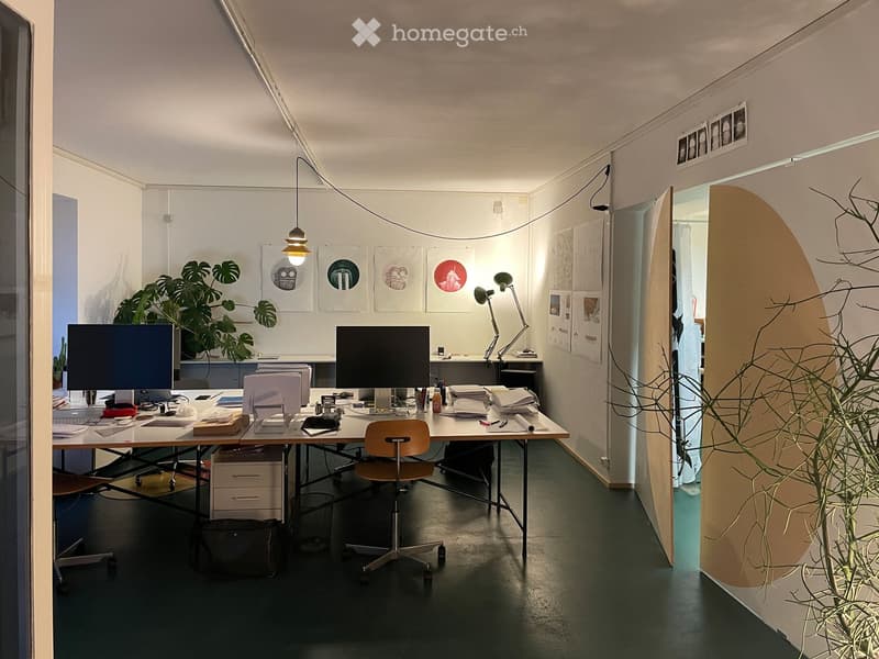 Modern office space with a large desk, two computer monitors, a chair, a lamp, and several framed artworks on the wall. There is a plant in the corner and a cabinet with drawers.