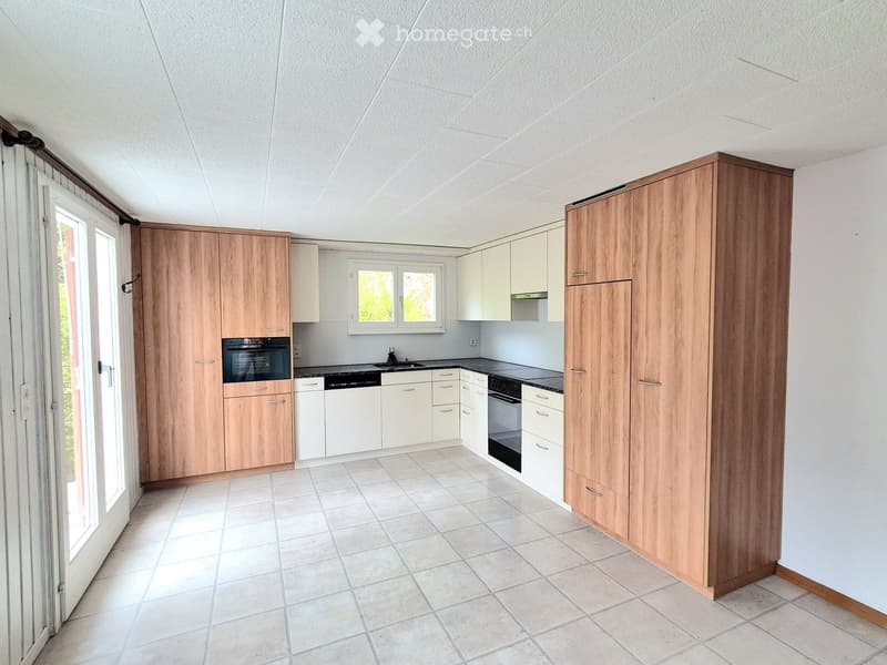 Modern kitchen with light wooden cabinets, white countertops, and built-in appliances including an oven, sink, and dishwasher. The space features tiled flooring and large windows allowing natural light.