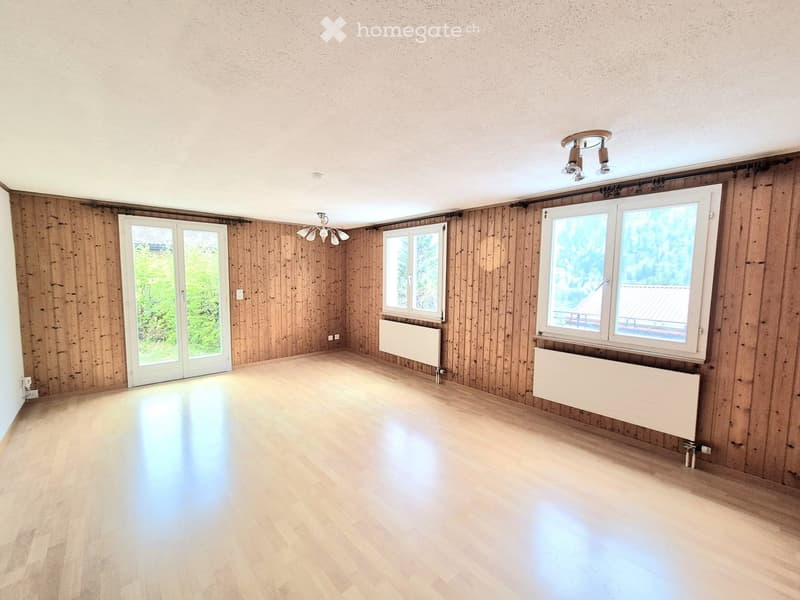 Empty room with wooden panel walls, light wooden flooring, large windows, and modern ceiling lights.