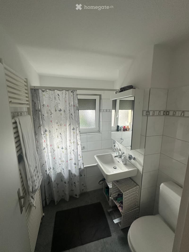 Bathroom with a bathtub, sink, toilet, mirror, radiator, towel, floral shower curtain, wicker basket, and a dark rug on the floor.