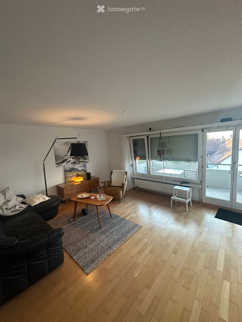 Modern living room with wooden flooring, large windows, and a balcony access. Features a black sectional sofa, coffee table, armchair, side table, floor lamp, and a picture frame on the wall.