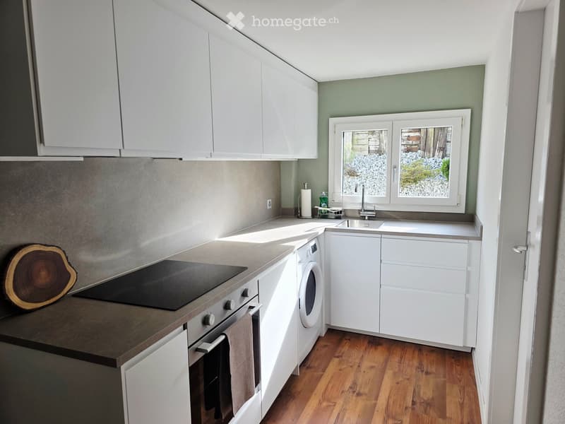 Modern kitchen with white cabinets, stainless steel appliances, and a wooden floor. Features an oven, washing machine, sink, and countertop.