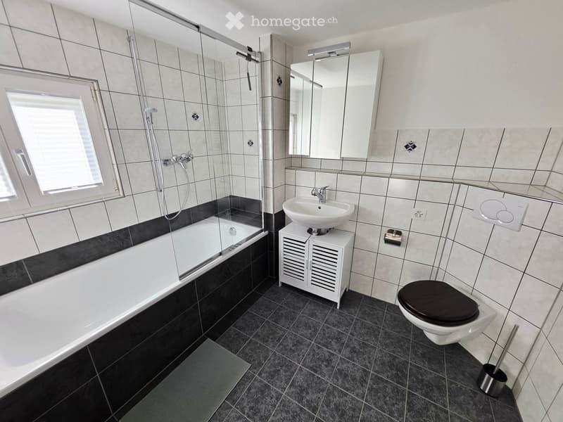 Modern bathroom with a bathtub, sink, toilet, and mirror. Features tiled walls and floor, a glass-enclosed shower area, and a small cabinet under the sink.